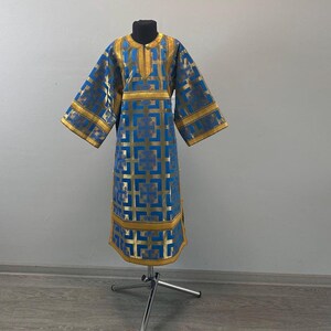 May include: A blue and gold liturgical garment with a square pattern design. The garment has gold trim on the sleeves, neckline, and hem. The garment is displayed on a black mannequin head and a silver stand. The logo "Ekklesia Church clothes" is visible.