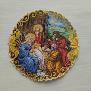Może przedstawiać: Okrągła naszywka haftowana przedstawiająca scenę narodzin. Naszywka przedstawia Marię, Józefa, małego Jezusa i inne postacie w kolorowym, szczegółowym wzorze. Naszywka jest otoczona złotą ramką z misternymi wzorami kwiatowymi.