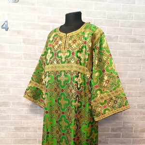 May include: A green and gold liturgical vestment with a cross pattern. The vestment has a round neckline and long sleeves. The fabric is brocade with a raised pattern.