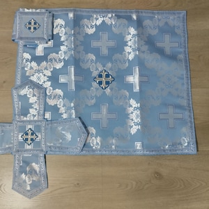 May include: A light blue altar set with embroidered crosses and floral patterns. The set includes a large rectangular piece, a smaller square piece, and a cross-shaped piece.