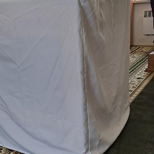 Op de afbeelding: Een groot, rechthoekig object bedekt met een lichtgrijze stof. Het staat op een houten voet, in een kamer met religieuze kunstwerken en een patroonkleed. De stof hangt over de randen.