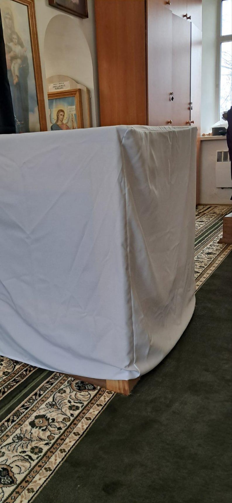 Op de afbeelding: Een groot, rechthoekig object bedekt met een lichtgrijze stof. Het staat op een houten voet, in een kamer met religieuze kunstwerken en een patroonkleed. De stof hangt over de randen.