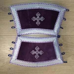 May include: Two purple velvet cushions with silver embroidered borders and a silver cross in the center. The cushions have blue ties on the sides.