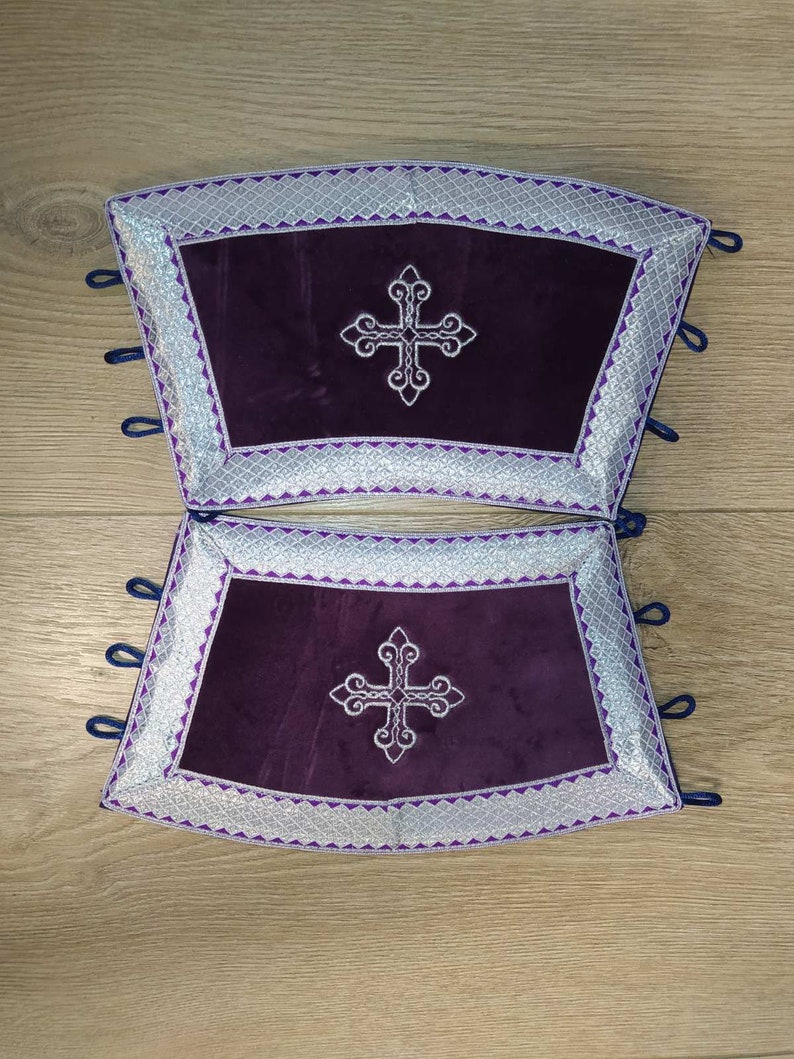 May include: Two purple velvet cushions with silver embroidered borders and a silver cross in the center. The cushions have blue ties on the sides.