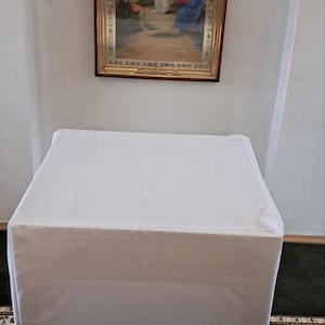 Op de afbeelding: Een witte, rechthoekige tafel bedekt met een witte doek, gepositioneerd voor een muur. Boven de tafel bevindt zich een religieuze icoon met een lijst, die een figuur met engelen afbeeldt.