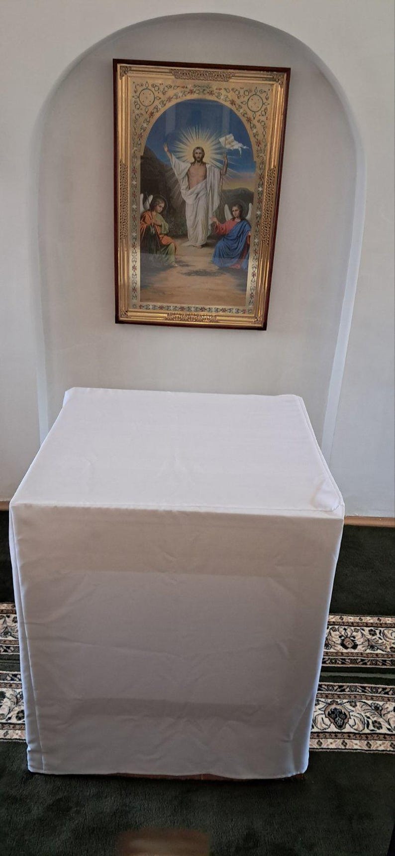 Op de afbeelding: Een witte, rechthoekige tafel bedekt met een witte doek, gepositioneerd voor een muur. Boven de tafel bevindt zich een religieuze icoon met een lijst, die een figuur met engelen afbeeldt.
