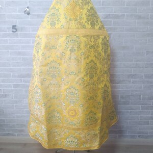 May include: A yellow liturgical vestment with a gold embroidered cross and floral pattern. The vestment is displayed on a metal stand.