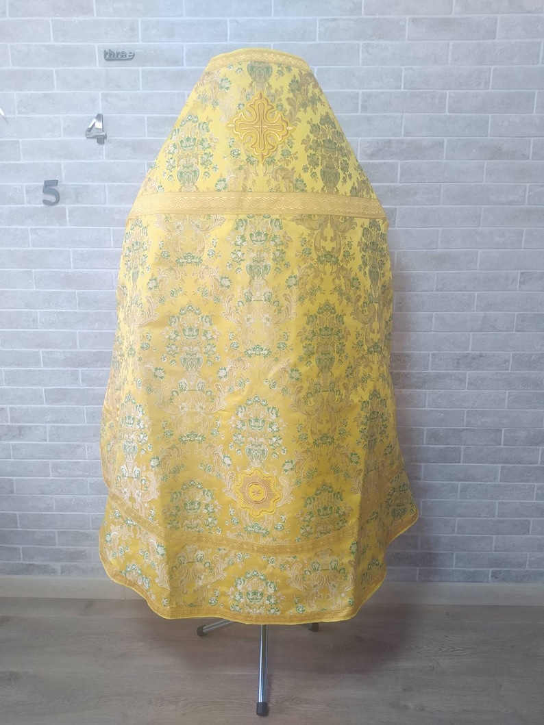 May include: A yellow liturgical vestment with a gold embroidered cross and floral pattern. The vestment is displayed on a metal stand.