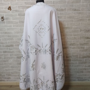 May include: White liturgical vestment with silver embroidery of wheat stalks, grapes, and a cross. The vestment is on a mannequin against a white brick wall.
