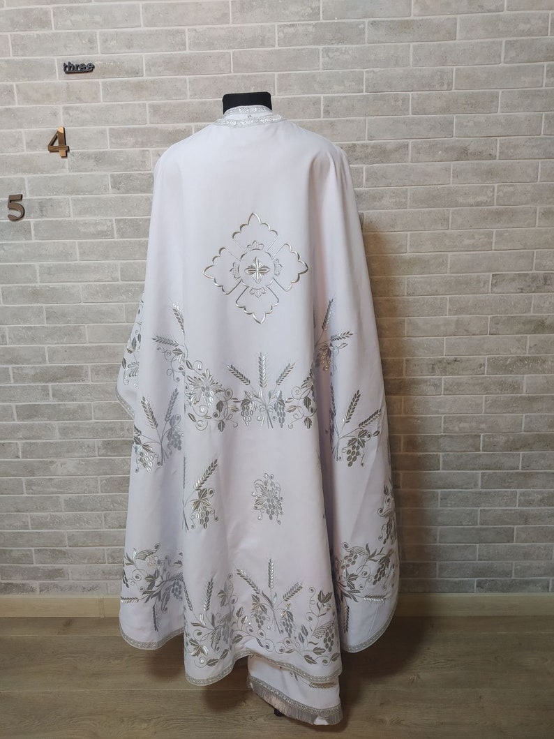 May include: White liturgical vestment with silver embroidery of wheat stalks, grapes, and a cross. The vestment is on a mannequin against a white brick wall.