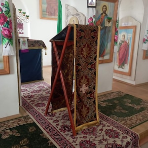 May include: A wooden lectern with a red and gold patterned fabric draped over it. The lectern is standing on a patterned rug in a room with white walls and religious artwork.