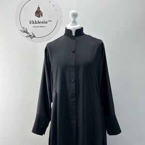 May include: A long, black church garment with a mandarin collar and button-down front. The sleeves are long with buttoned cuffs. The garment is displayed on a white mannequin head. The logo for Ekklesia Church clothes is visible in the background.