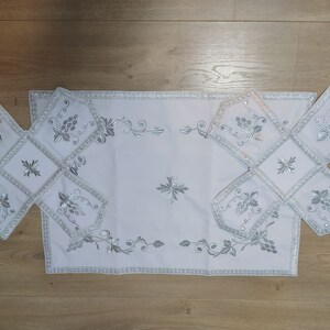May include: White embroidered table linens with a silver floral design. The linens are arranged in a cross shape, with a larger rectangular piece in the center.
