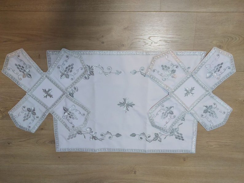 May include: White embroidered table linens with a silver floral design. The linens are arranged in a cross shape, with a larger rectangular piece in the center.