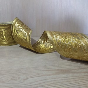 May include: Gold ribbon with a floral pattern. The ribbon is rolled up and has a shiny finish.