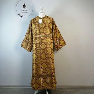 May include: A gold and burgundy liturgical vestment with an ornate floral pattern. The long-sleeved garment is displayed on a white mannequin. The brand logo, "Ekklesia Church clothes," is visible in the background.
