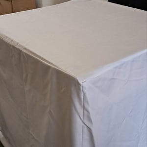 Op de afbeelding: Een witte rechthoekige tafel bedekt met een witte doek. De doek bedekt de randen van de tafel, waardoor een strakke, minimalistische esthetiek ontstaat. De tafel staat in een kamer met andere objecten op de achtergrond.