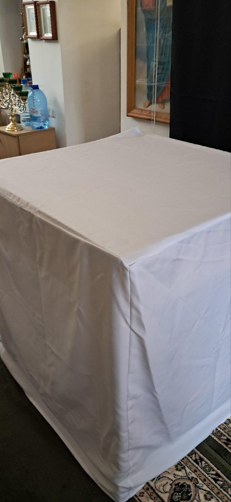 Op de afbeelding: Een witte rechthoekige tafel bedekt met een witte doek. De doek bedekt de randen van de tafel, waardoor een strakke, minimalistische esthetiek ontstaat. De tafel staat in een kamer met andere objecten op de achtergrond.