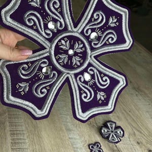 May include: A large purple and silver embroidered cross with intricate details and rhinestones. Two smaller crosses are also visible in the background.
