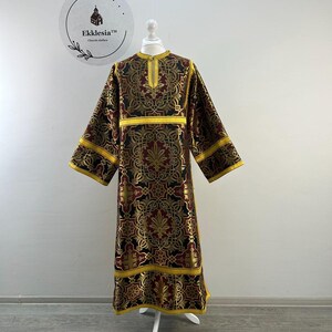 May include: A long, ornate church garment with a high collar, long sleeves, and a floor-length hem. The fabric is a rich combination of gold, burgundy, and black, with intricate floral and geometric patterns. Yellow trim accents the sleeves and hem. The garment is displayed on a white mannequin.