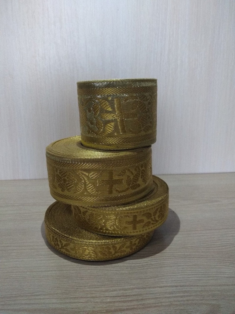 May include: Four spools of gold ribbon with a cross and floral pattern. The ribbon is stacked on top of each other.