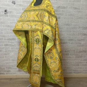 May include: A yellow and green patterned liturgical vestment with gold accents. The vestment is displayed on a stand against a brick wall.