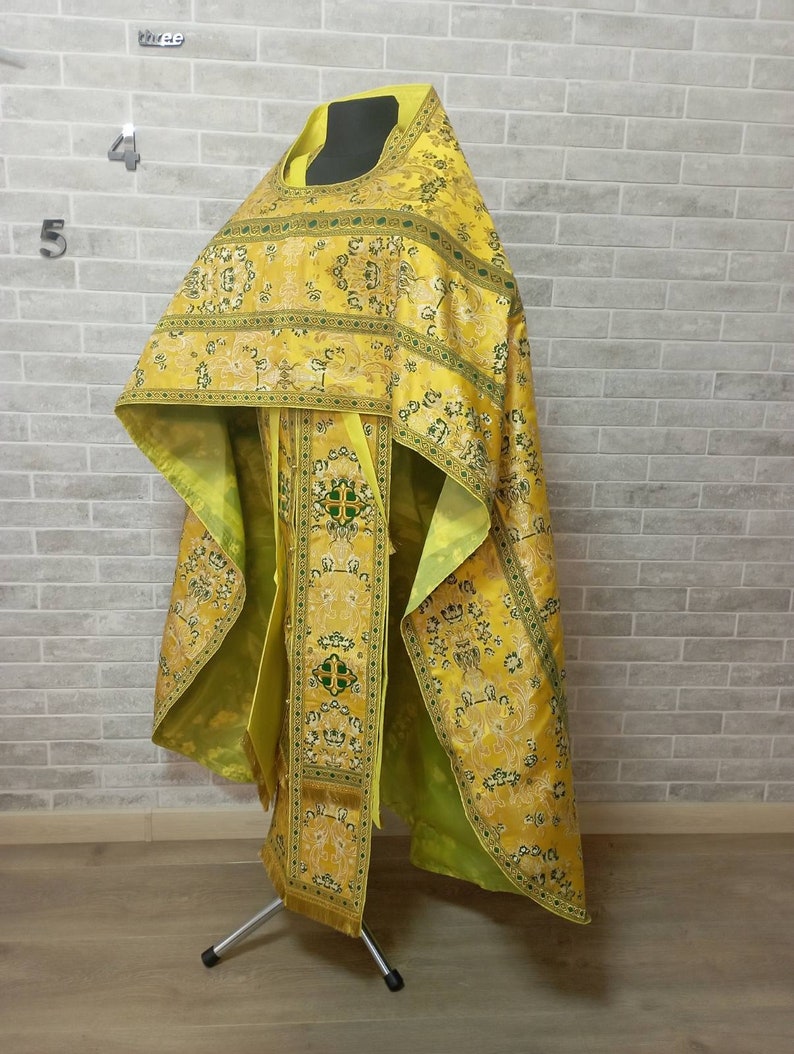 May include: A yellow and green patterned liturgical vestment with gold accents. The vestment is displayed on a stand against a brick wall.