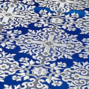 Pu&ograve; includere: Primo piano di un tessuto blu e argento con un motivo floreale ornato. Il design presenta viticci, fiori e forme geometriche argentate su uno sfondo blu intenso. Il tessuto ha un aspetto strutturato.