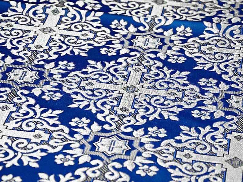 Pu&ograve; includere: Primo piano di un tessuto blu e argento con un motivo floreale ornato. Il design presenta viticci, fiori e forme geometriche argentate su uno sfondo blu intenso. Il tessuto ha un aspetto strutturato.