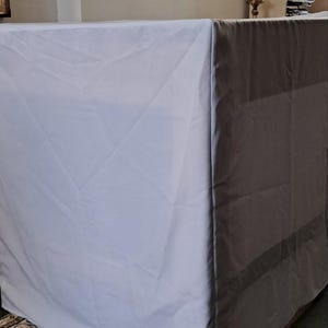 Op de afbeelding: Een witte rechthoekige tafel bedekt met een witte doek. De tafel staat in een kamer met een schilderij en een boekenplank. De tafel wordt waarschijnlijk gebruikt voor een religieuze ceremonie.