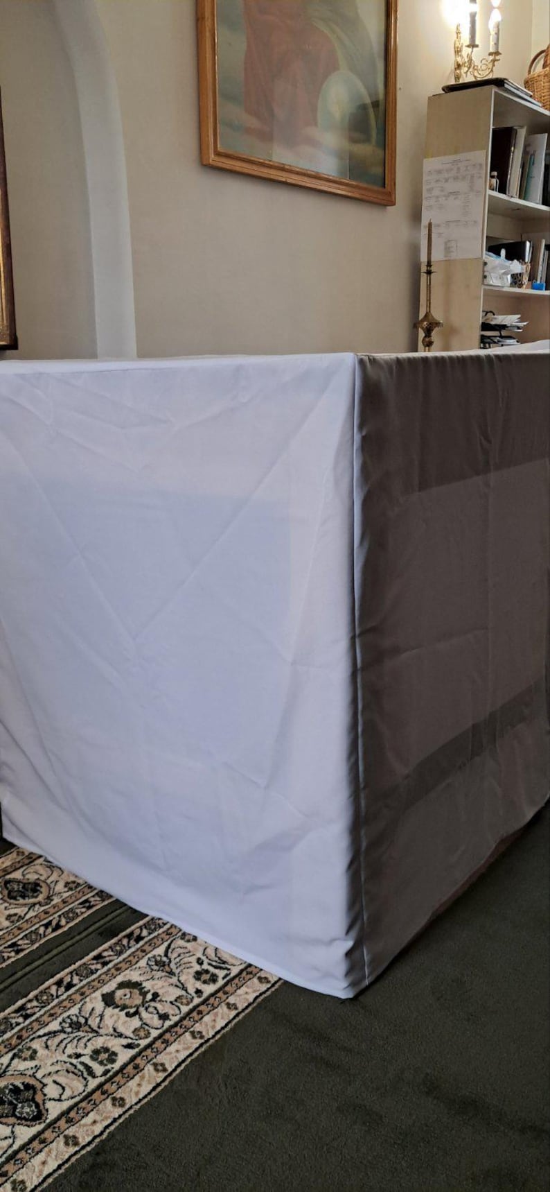 Op de afbeelding: Een witte rechthoekige tafel bedekt met een witte doek. De tafel staat in een kamer met een schilderij en een boekenplank. De tafel wordt waarschijnlijk gebruikt voor een religieuze ceremonie.