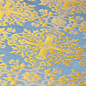 Pu&ograve; includere: Primo piano di un tessuto blu con un motivo floreale dorato. Il design include croci e volute ornate. Il tessuto ha una consistenza morbida e vellutata e una lucentezza sottile, creando un aspetto lussuoso.