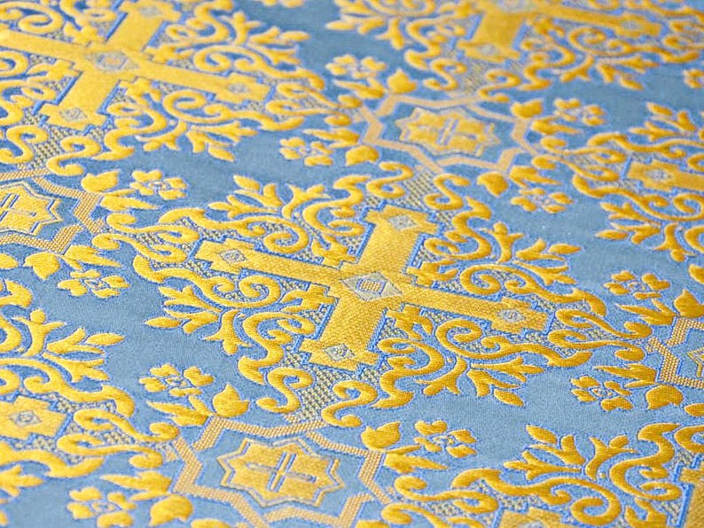 Pu&ograve; includere: Primo piano di un tessuto blu con un motivo floreale dorato. Il design include croci e volute ornate. Il tessuto ha una consistenza morbida e vellutata e una lucentezza sottile, creando un aspetto lussuoso.