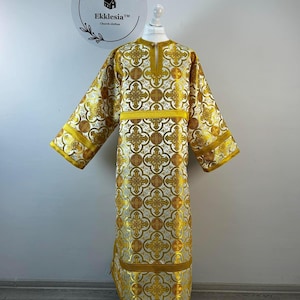May include: A long, ornate liturgical garment with a gold and white pattern, displayed on a mannequin. The garment features a repeating cross design and gold trim. The text "Ekklesia Church clothes" is visible in the background.