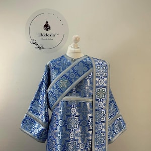 May include: A blue and silver liturgical vestment featuring a repeating geometric pattern with cross motifs. The garment includes a wide diagonal stole draped over the shoulder, long sleeves, and silver trim along the edges and neckline.