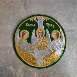 Puede incluir: Un parche circular verde bordado con hilo dorado y plateado que representa a tres ángeles con alas, sentados alrededor de una mesa. El texto "Святая Троица" está bordado en hilo dorado en el parche.