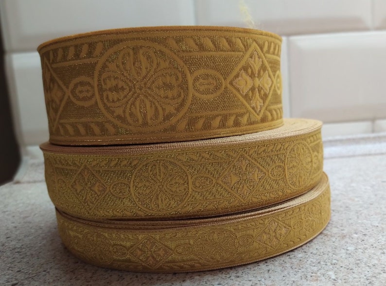 Puede incluir: Tres galones de tela de color dorado con intrincados patrones en relieve. Los galones est&aacute;n apilados uno encima del otro, creando un efecto de capas.