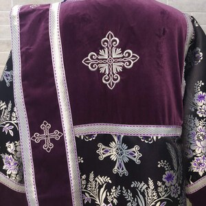 May include: A purple liturgical vestment with silver embroidery of crosses and floral patterns. The vestment has a long, flowing design and is made of a luxurious fabric.