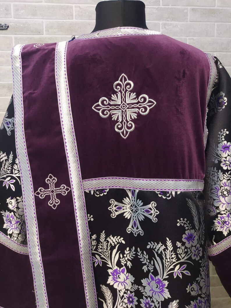 May include: A purple liturgical vestment with silver embroidery of crosses and floral patterns. The vestment has a long, flowing design and is made of a luxurious fabric.