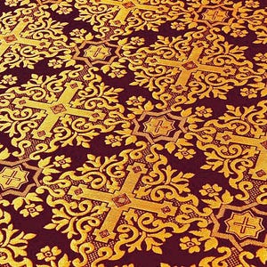 Pu&ograve; includere: Primo piano di un tessuto bordeaux con un motivo dorato ripetuto. Il design presenta croci ornate ed elementi floreali, creando un aspetto ricco e strutturato. I dettagli intricati del tessuto suggeriscono un uso decorativo o cerimoniale.
