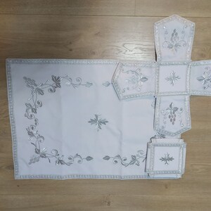 May include: A white embroidered altar cloth set with a cross and grapevine designs. The set includes a large rectangular cloth, a smaller square cloth, and four smaller rectangular cloths.