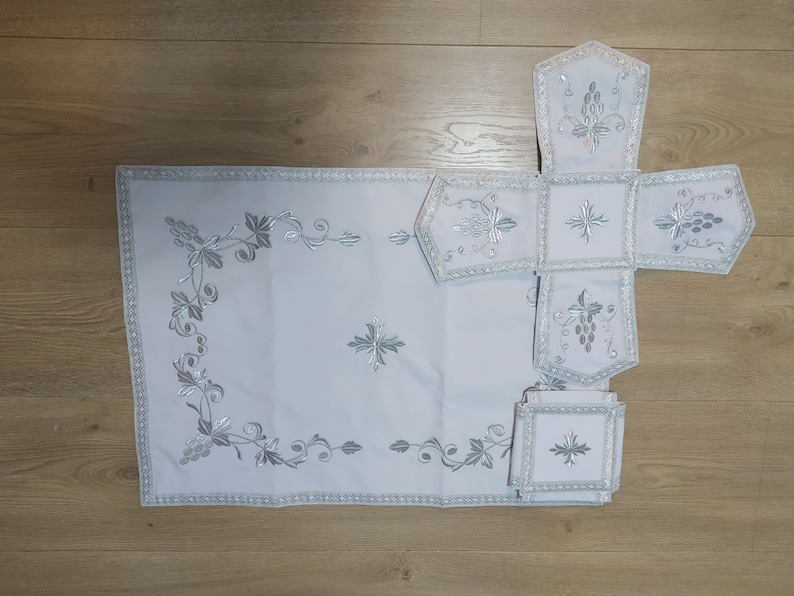 May include: A white embroidered altar cloth set with a cross and grapevine designs. The set includes a large rectangular cloth, a smaller square cloth, and four smaller rectangular cloths.