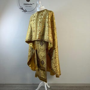 May include: A gold-colored liturgical vestment set featuring a repeating pattern of crosses within circular motifs. The garment includes a cape-like upper section and a long, narrow stole with decorative fringe and embroidered cross details.