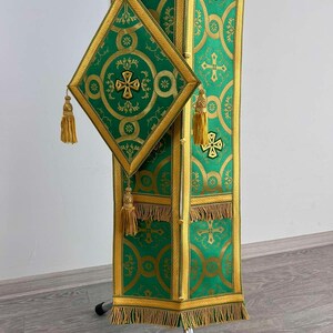 May include: A green and gold liturgical stole with a matching green and gold square pocket. The stole has a fringe trim and the pocket has a tassel.