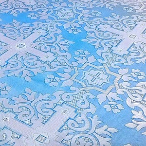 Pu&ograve; includere: Primo piano di un tessuto azzurro con un motivo ripetuto di croci bianche, motivi floreali e volute ornate. Il tessuto ha una leggera lucentezza, che suggerisce un materiale satinato o damascato, e viene probabilmente utilizzato per paramenti religiosi o scopi decorativi.