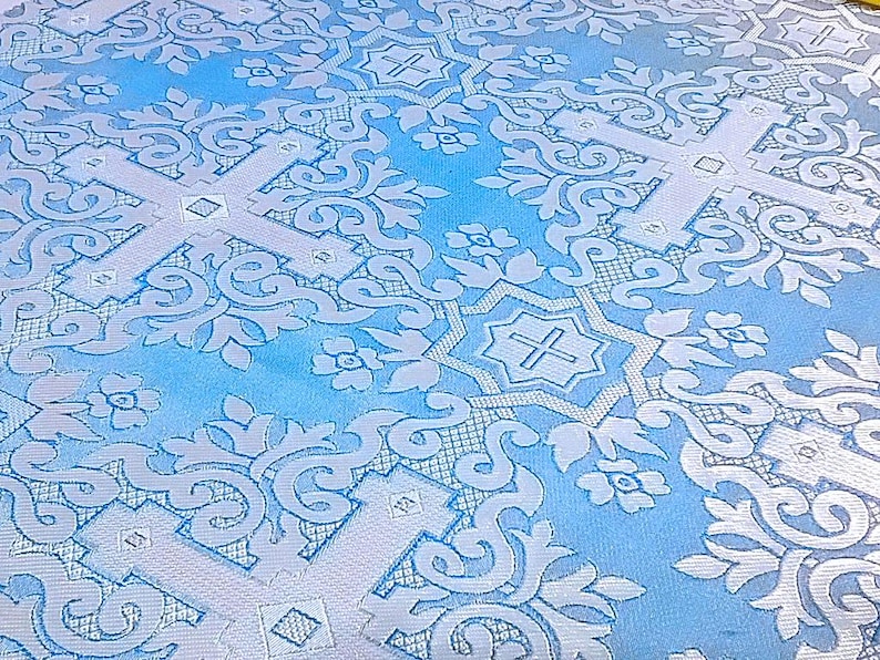 Pu&ograve; includere: Primo piano di un tessuto azzurro con un motivo ripetuto di croci bianche, motivi floreali e volute ornate. Il tessuto ha una leggera lucentezza, che suggerisce un materiale satinato o damascato, e viene probabilmente utilizzato per paramenti religiosi o scopi decorativi.