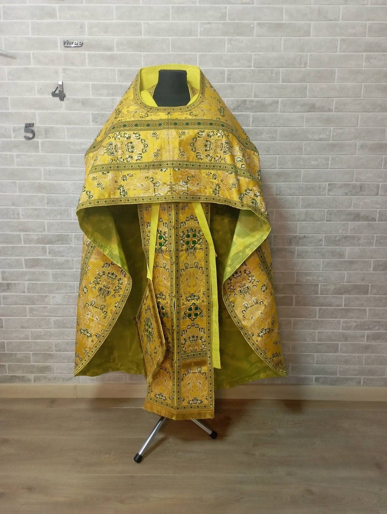 May include: A yellow and green liturgical vestment with intricate embroidery and gold accents. The vestment is displayed on a stand against a brick wall.