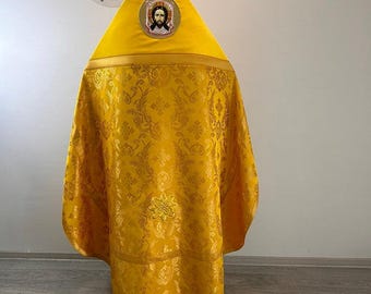 May include: A bright yellow liturgical vestment featuring a pointed hood with a circular icon of Jesus Christ. The garment is crafted from a patterned fabric with intricate floral and cross motifs, finished with a solid yellow band at the top.