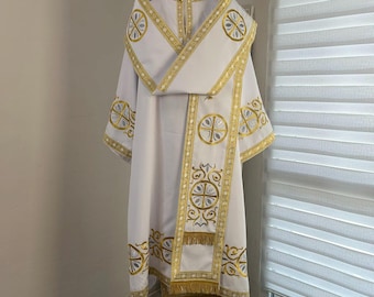 May include: A white liturgical vestment featuring intricate gold embroidery and circular cross motifs. The garment includes a long stole with gold fringe at the bottom, a wide collar, and decorative trim along the edges, displayed on a mannequin.