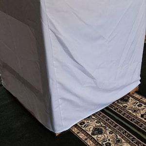 Op de afbeelding: Een witte tafelhoes over een vierkante tafel. De tafel staat op een patroonkleed met een donkergroene achtergrond. De stof lijkt glad en schoon.